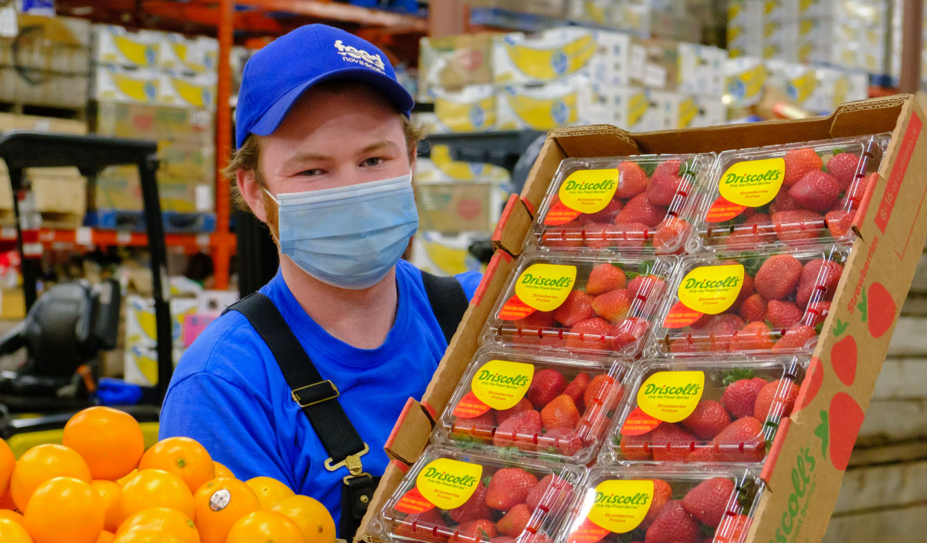 Person standing in a warehouse, wearing a Feed Nova Scotia hat and holding fresh strawberries and oranges