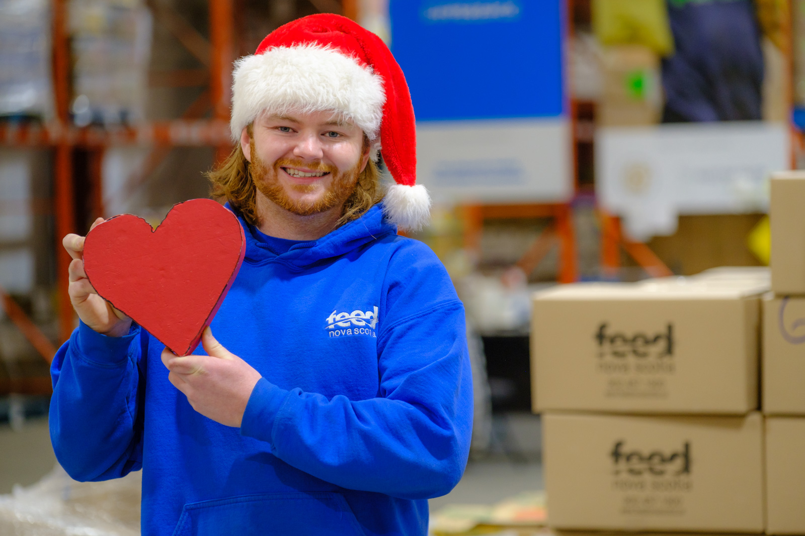 Person standing in a warehouse wearing a Santa hat and smiling and holding a small red heart