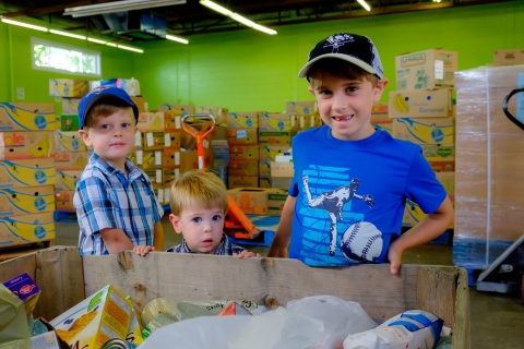 Three young boys standing in front of a box filled with food